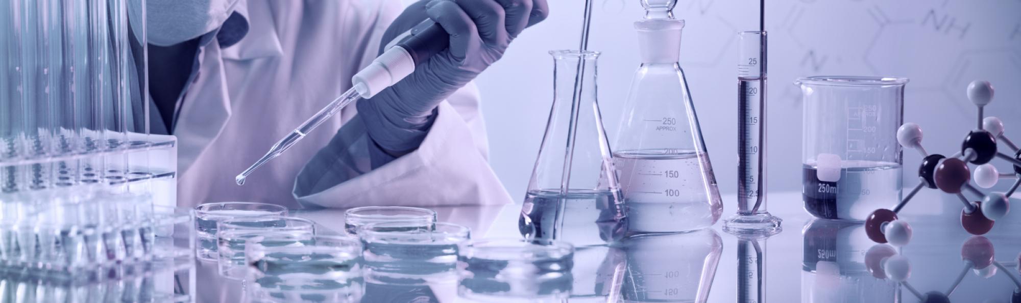 a scientist using a pipette surrounded by beakers, petri dishes and a model of an atom