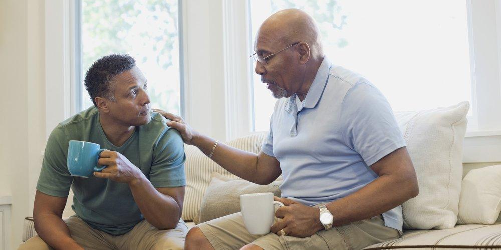 An African American elder father and middle aged son drinking coffee and talking.