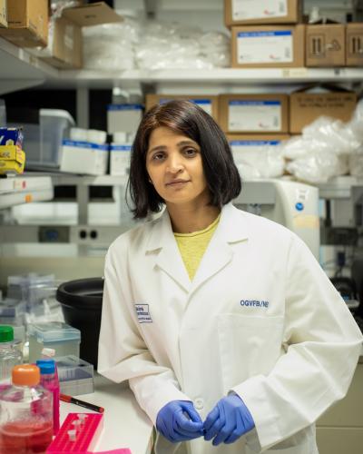 Woman wearing a white lab coat and blue gloves in a lab setting with bottles and boxes behind her.