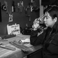 A woman, putting on goggles, sits at a workbench with tools. Image in black and white.